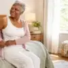 Senior woman putting on a cardigan while sitting on the bed